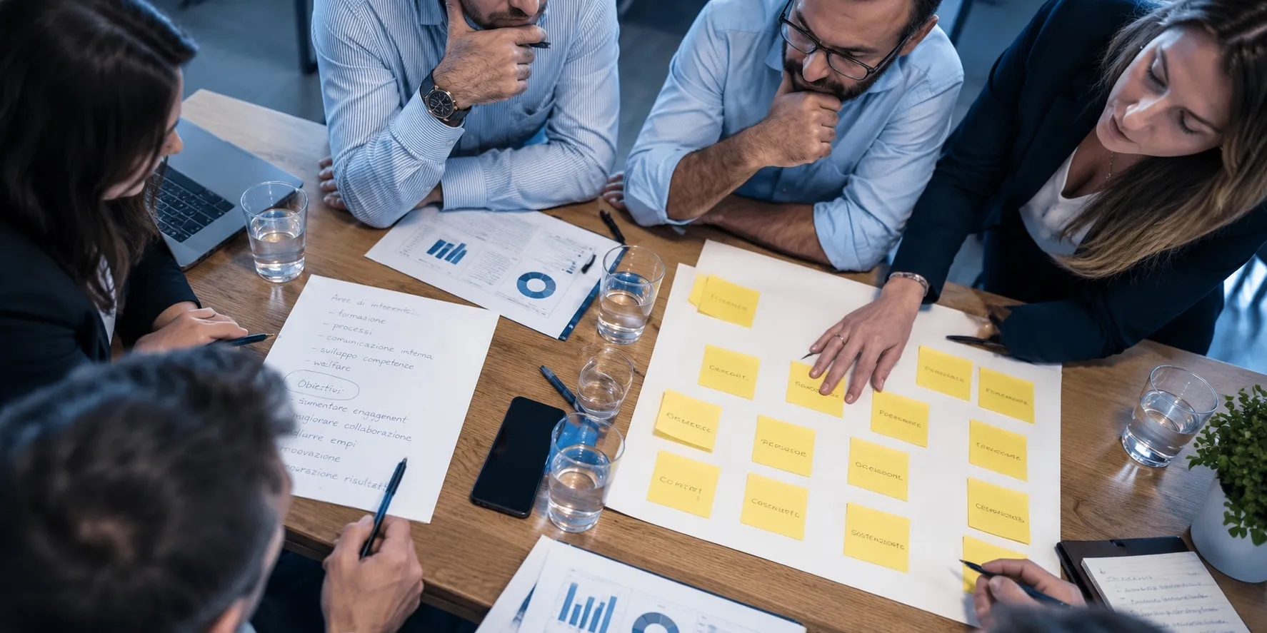 Colleghi attorno a un tavolo, visti dall’alto, al lavoro su post-it, grafici e appunti.